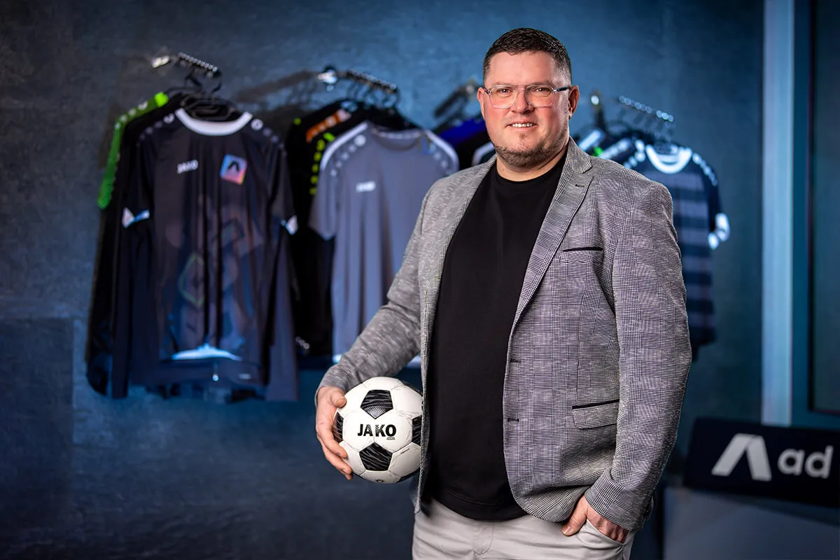 Geschäftsführer mit Fußball vor einer Wand mit Sporttrikots